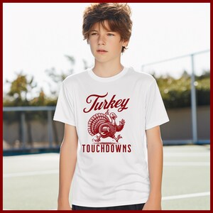 Op de afbeelding: Wit T-shirt met een rode afbeelding van een kalkoen met een voetbal en de woorden "Turkey & Touchdowns". Het shirt heeft korte mouwen en wordt door een persoon gedragen.
