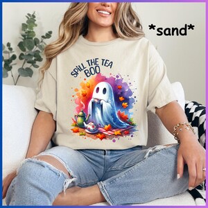 Pu&ograve; includere: Una maglietta beige con una grafica di un fantasma che tiene una tazza di t&egrave; e il testo "Spill the Tea Boo". Il fantasma &egrave; seduto su una coperta da picnic con foglie autunnali e una teiera.