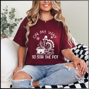Puede incluir: Una camiseta color granate con un gráfico blanco de un pavo que lleva un sombrero de peregrino y remueve una olla. El texto de la camiseta dice "On my way to stir the pot."
