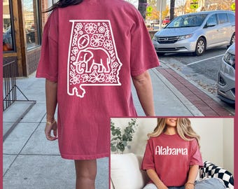 May include: A maroon t-shirt featuring a white outline of the state of Alabama, filled with floral and elephant designs. The back of the shirt displays the state outline with a football and elephant. The word "Alabama" is printed on the front of the shirt.