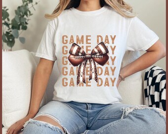 May include: White t-shirt with a football-themed graphic. The design features a football-shaped bow with brown and white stripes and the words "GAME DAY" in a repeating pattern. The shirt is worn with blue jeans.