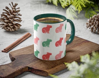 Può includere: Una tazza in ceramica bianca con un bordo e un manico verdi. La tazza presenta un motivo ripetuto di capibara rossi e verdi. La tazza è piena di caffè e dal bordo sale vapore.