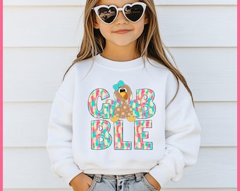 Op de afbeelding: Een witte sweatshirt met het woord "GOBBLE" in kleurrijke, patroonletters. Een cartoon kalkoen met een blauwe strik is gecentreerd boven de "B" in "GOBBLE". De sweatshirt wordt gedragen door een jong persoon met een hartvormige zonnebril.