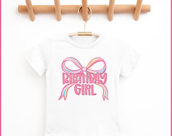 May include: White t-shirt with a colorful bow design and the words "BIRTHDAY GIRL" in pink. The shirt is hanging on a wooden rack with a gold hanger. The bow has rainbow-colored accents.