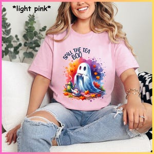 Pu&ograve; includere: Una maglietta rosa chiaro con un'immagine di un fantasma che tiene una tazza di t&egrave; e il testo "Spill the Tea Boo".