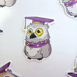 May include: A sticker featuring a cartoon owl wearing a purple graduation cap, gold-rimmed glasses, and a purple scarf with yellow stars. The owl has gray and white feathers and a black beak. The sticker is on a white background.