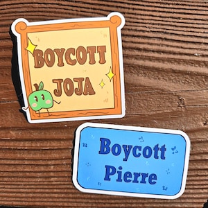 Könnte beinhalten: Zwei Aufkleber. Einer zeigt die Worte "BOYCOTT JOJA" in Braun auf beigem Hintergrund, mit einem Cartoon-Apfel. Der andere sagt "Boycott Pierre" in Blau auf blauem Hintergrund, mit kleinen dekorativen Elementen.