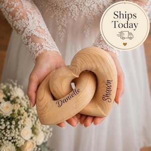 May include: Two wooden heart-shaped objects interlocked, held in hands. One heart is engraved with "Danielle," the other with "Shawn." The wood has a light, natural finish. The background includes a bouquet of white flowers and a wedding dress.