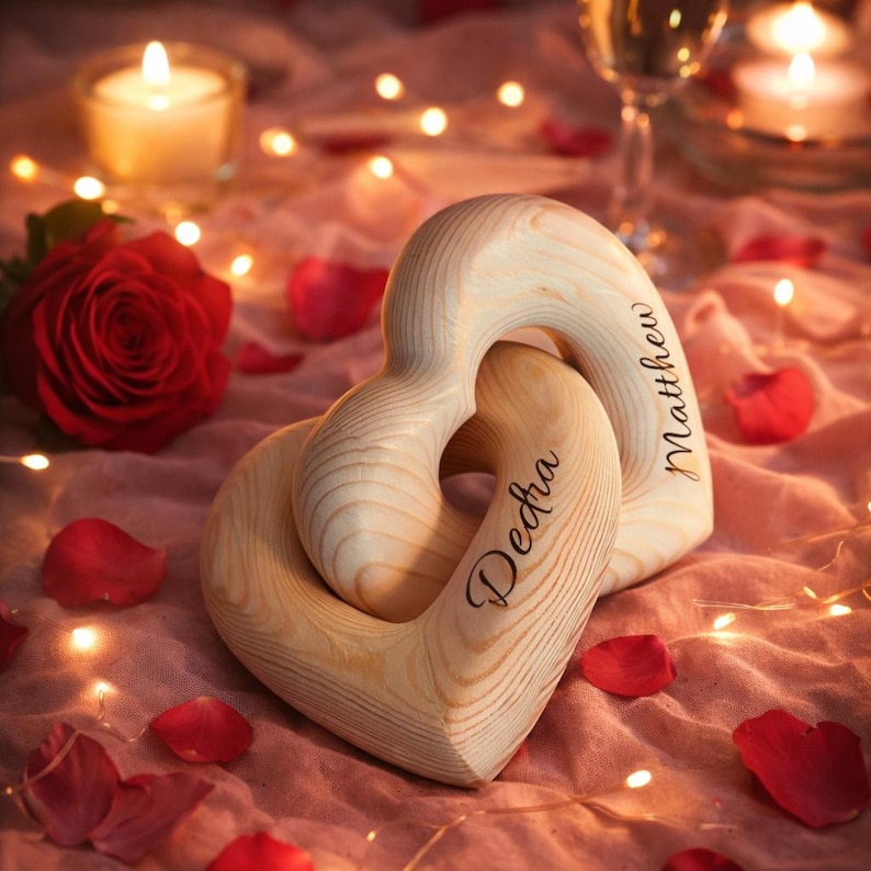 May include: Two interlocking wooden heart-shaped ornaments. One heart is engraved with the name "Dedra," and the other with "Matthew." The ornaments are light-colored wood, resting on a pink surface with rose petals and a red rose.
