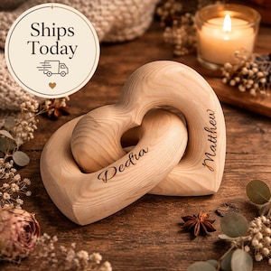Puede incluir: Dos adornos entrelazados de madera en forma de corazón con los nombres "Dedra" y "Matthew" grabados. Los adornos de color marrón claro descansan sobre una superficie de madera. También se ve una vela encendida y el texto "Ships Today".
