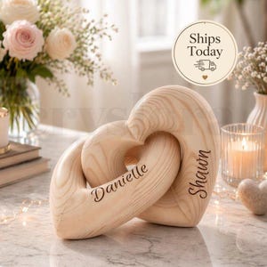 May include: Wooden heart sculpture with interlocking design. The sculpture is light brown with the names "Danielle" and "Shawn" engraved on the sides. The background includes flowers, candles, and the text "Ships Today".