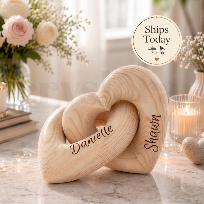 K&ouml;nnte beinhalten: Holzherzskulptur mit ineinandergreifendem Design. Die Skulptur ist hellbraun und tr&auml;gt die Namen "Danielle" und "Shawn" auf den Seiten eingraviert. Der Hintergrund umfasst Blumen, Kerzen und den Text "Ships Today".