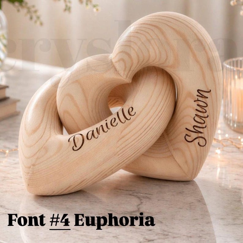K&ouml;nnte beinhalten: Zwei ineinandergreifende Holzherzen mit den eingravierten Namen "Danielle" und "Shawn". Die Herzen sind aus hellem Holz und ineinander verschlungen. Der Text "Font #4 Euphoria" steht unten.