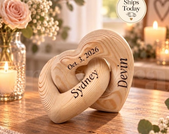May include: Two interlocking wooden hearts with a natural grain finish. The hearts are engraved with the names Sydney and Devin, along with the date Oct. 3, 2026. The object features a smooth, curved design and is displayed on a wooden surface.