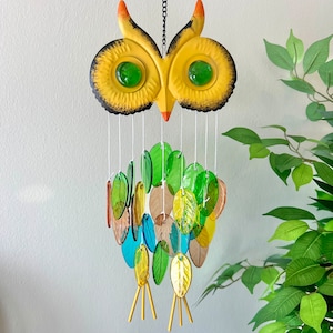 May include: A yellow owl wind chime with green, blue, and brown glass pieces hanging from it. The owl has black eyes and a black outline.