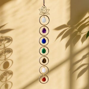 May include: A decorative chakra wall hanging featuring seven teardrop-shaped gemstones in various colors, suspended within gold-colored rings. The top features a lotus flower design. The gemstones include white, purple, blue, green, brown, white, and red.