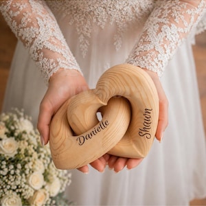 May include: Two interlocking wooden hearts, engraved with the names "Danielle" and "Shawn", are held in front of a white dress. The hearts are made of light-colored wood, with a natural grain pattern. The names are written in a cursive font.