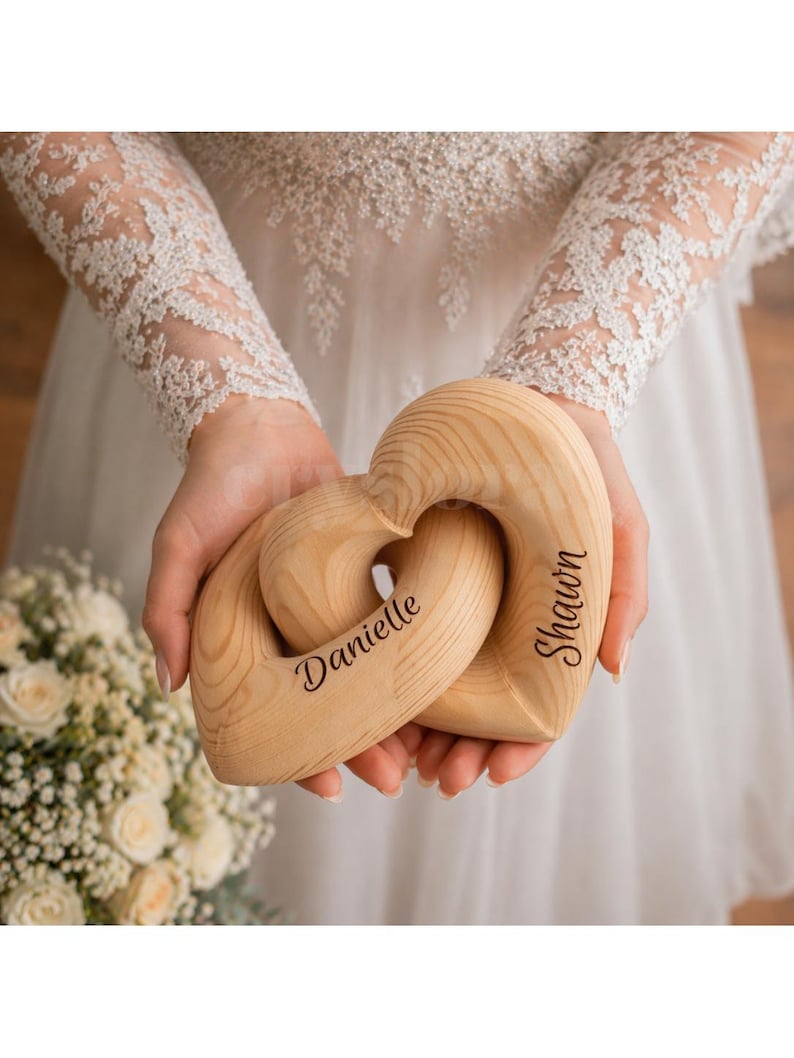 May include: Two interlocking wooden hearts, engraved with the names "Danielle" and "Shawn", are held in front of a white dress. The hearts are made of light-colored wood, with a natural grain pattern. The names are written in a cursive font.