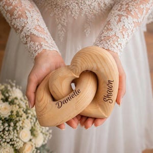 May include: Two interlocking wooden heart-shaped sculptures made of light-colored wood with visible grain patterns. The left heart is engraved with the name Danielle in a dark script font, and the right heart is engraved with the name Shawn.