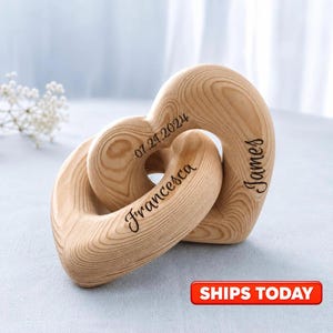 May include: Two interlocking light-colored wooden heart shapes with a visible grain pattern. The wood is engraved with the name Francesca, the name James, and the date 07.21.2024 in a dark script font. The hearts are connected to form a single piece.