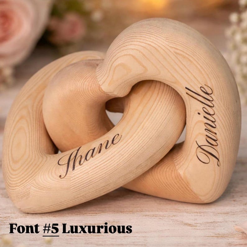 May include: Interlocking wooden heart-shaped rings with the names "Shane" and "Danielle" engraved in a cursive font. The rings are made of light-colored wood and are interlocked. The text "Font #5 Luxurious" is at the bottom.