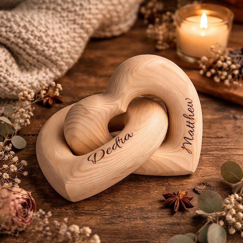 May include: Two interlocking wooden hearts with the names "Dedra" and "Matthew" engraved on them. The hearts are light-colored wood and are intertwined. The background includes a candle, flowers, and a wooden surface.