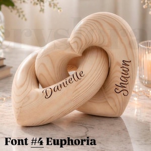 May include: A wooden sculpture of two intertwined hearts. One heart is engraved with the name "Danielle", the other with "Shawn". The sculpture is made of light-colored wood and features the text "Font #4 Euphoria" at the bottom.
