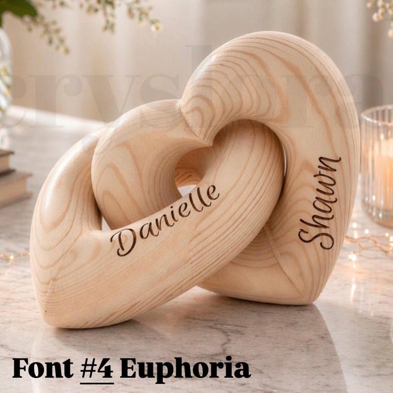 May include: A wooden sculpture of two intertwined hearts. One heart is engraved with the name "Danielle", the other with "Shawn". The sculpture is made of light-colored wood and features the text "Font #4 Euphoria" at the bottom.