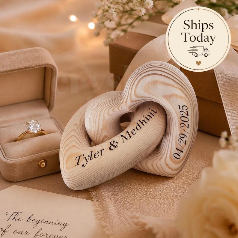 May include: A wooden heart-shaped wedding keepsake with the names "Tyler & Medhini" and the date "04/29/2025" engraved on it. A gold engagement ring in a velvet box is also visible. The background includes a gift box and flowers.