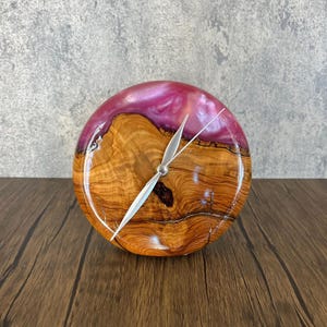 Puede incluir: Un reloj redondo de madera con una incrustación de resina rosa. La esfera del reloj tiene agujas plateadas y está colocada sobre un fondo de veta de madera natural.