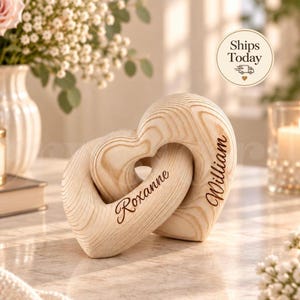 Puede incluir: Dos corazones de madera entrelazados con un acabado de veta de madera clara natural. El corazón izquierdo está grabado con el nombre Roxanne en letra cursiva, y el corazón derecho está grabado con el nombre William en letra cursiva a juego.