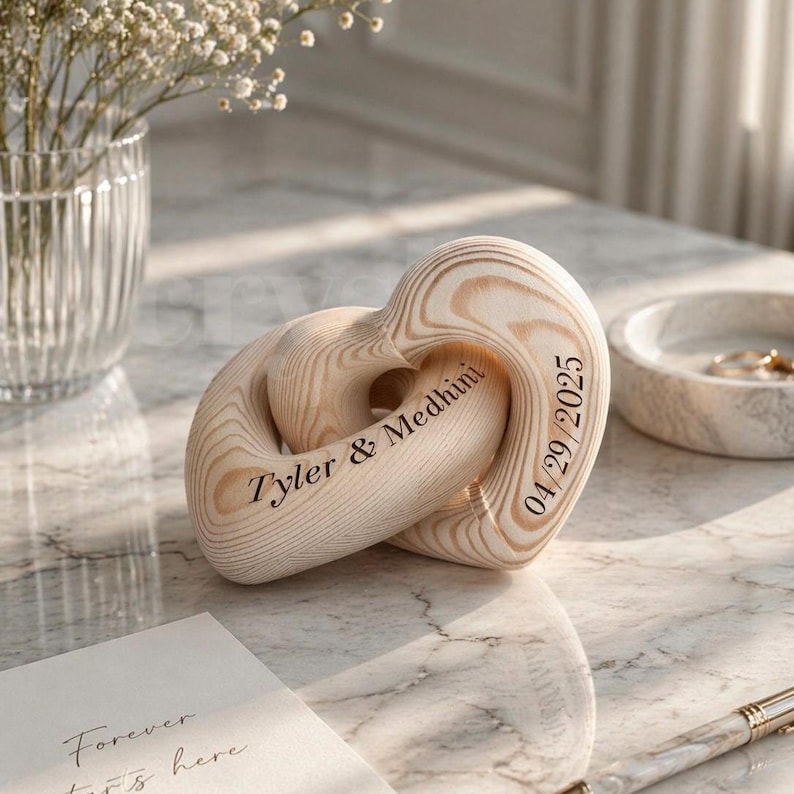 May include: A wooden heart-shaped sculpture with interlocking loops. The sculpture has a light wood grain pattern and features the names "Tyler & Medhini" and the date "04/29/2025" engraved on it. The sculpture is on a marble surface.