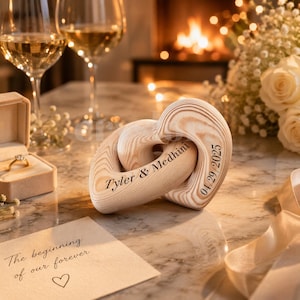 May include: A wooden heart-shaped sculpture with interlocking rings, engraved with the names "Tyler & Medhini" and the date "04/29/2025". The sculpture is light brown and sits on a reflective surface, with a ring box and a card that says "The beginning of our forever".
