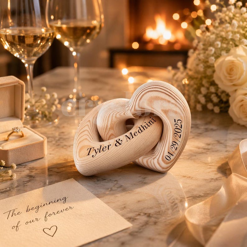 May include: A wooden heart-shaped sculpture with interlocking rings, engraved with the names "Tyler & Medhini" and the date "04/29/2025". The sculpture is light brown and sits on a reflective surface, with a ring box and a card that says "The beginning of our forever".