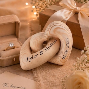 May include: A gold engagement ring in a velvet box next to two wooden heart-shaped ornaments. The ornaments are engraved with "Tyler & Medhini" and "04/29/2025". A gift box with a ribbon is in the background.