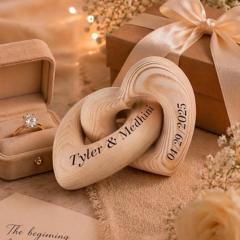 May include: A gold engagement ring in a velvet box next to two wooden heart-shaped ornaments. The ornaments are engraved with "Tyler & Medhini" and "04/29/2025". A gift box with a ribbon is in the background.