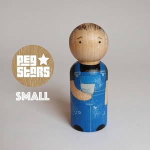 May include: A hand-painted wooden peg doll wearing blue overalls and holding a small cup. The doll has a brown painted face with black eyes and a brown painted hair.