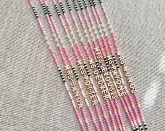 Puede incluir: Una colección de cordones con cuentas en rosa, blanco, negro y dorado. Cada cordón presenta un nombre en letras doradas. Los cordones tienen un cordón blanco y cierres dorados. Las cuentas están dispuestas en patrones.