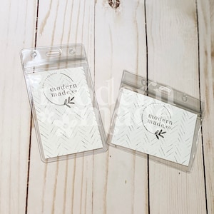 May include: Two clear plastic badge holders with a white card inside. The card has a black and white pattern and the text "modern made co." in a circle.