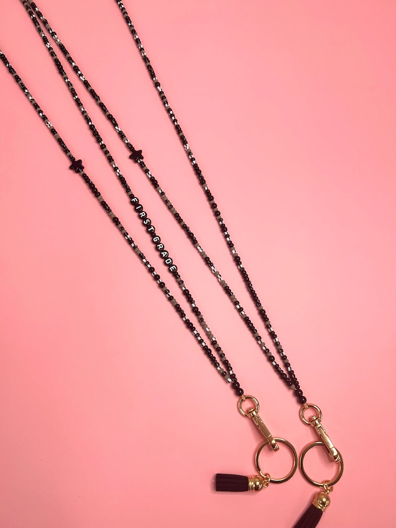 May include: Two black beaded necklaces with gold clasps and tassels. One necklace has the word "FIRST GRADE" spelled out in black beads. Both necklaces have a small black star charm.