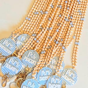 May include: A collection of teacher lanyards with wooden beads and blue star accents. Each lanyard features a round charm with a blue and white striped pattern and a teacher's name in white lettering. Gold-colored keychains and white tassels are attached.