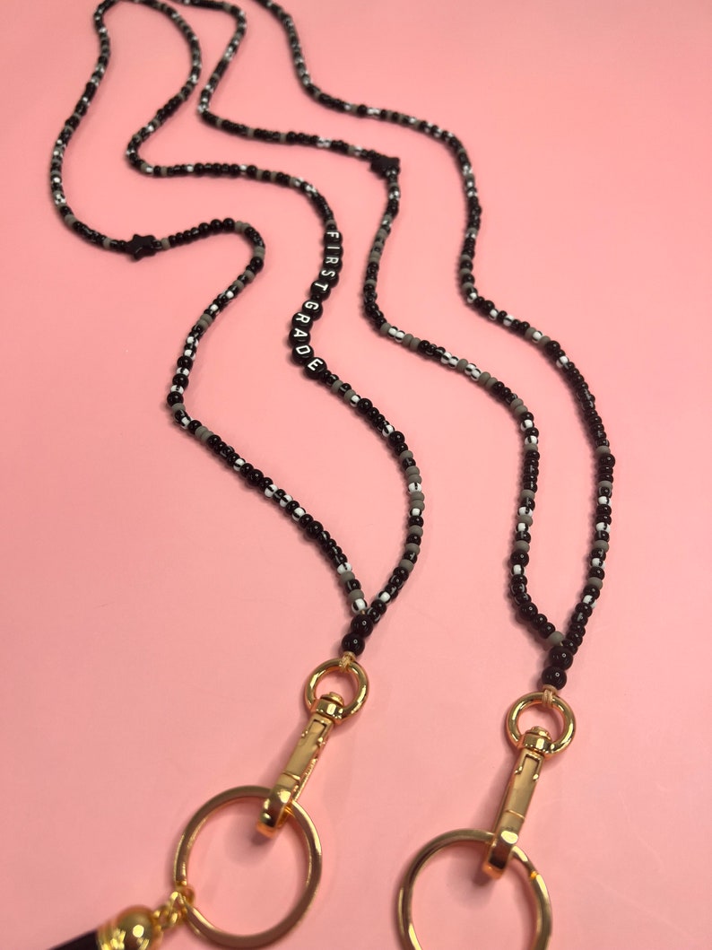 May include: Black and white beaded lanyard with gold clasps and a black beaded charm with the text "FIRST GRADE" in white letters.