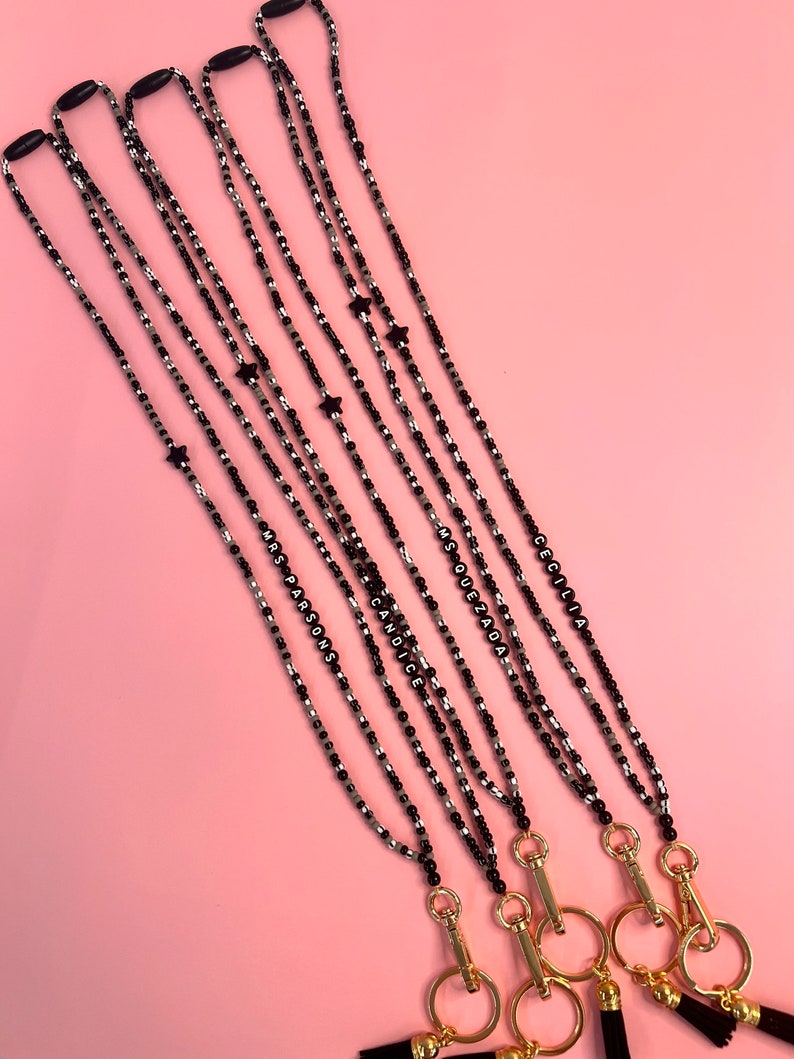May include: Five black and silver beaded lanyards with gold clasps and black tassels. Each lanyard has a black star charm and a name spelled out in black beads: Cecilia, Ms. Quezada, Candice, Ms. Parsons.