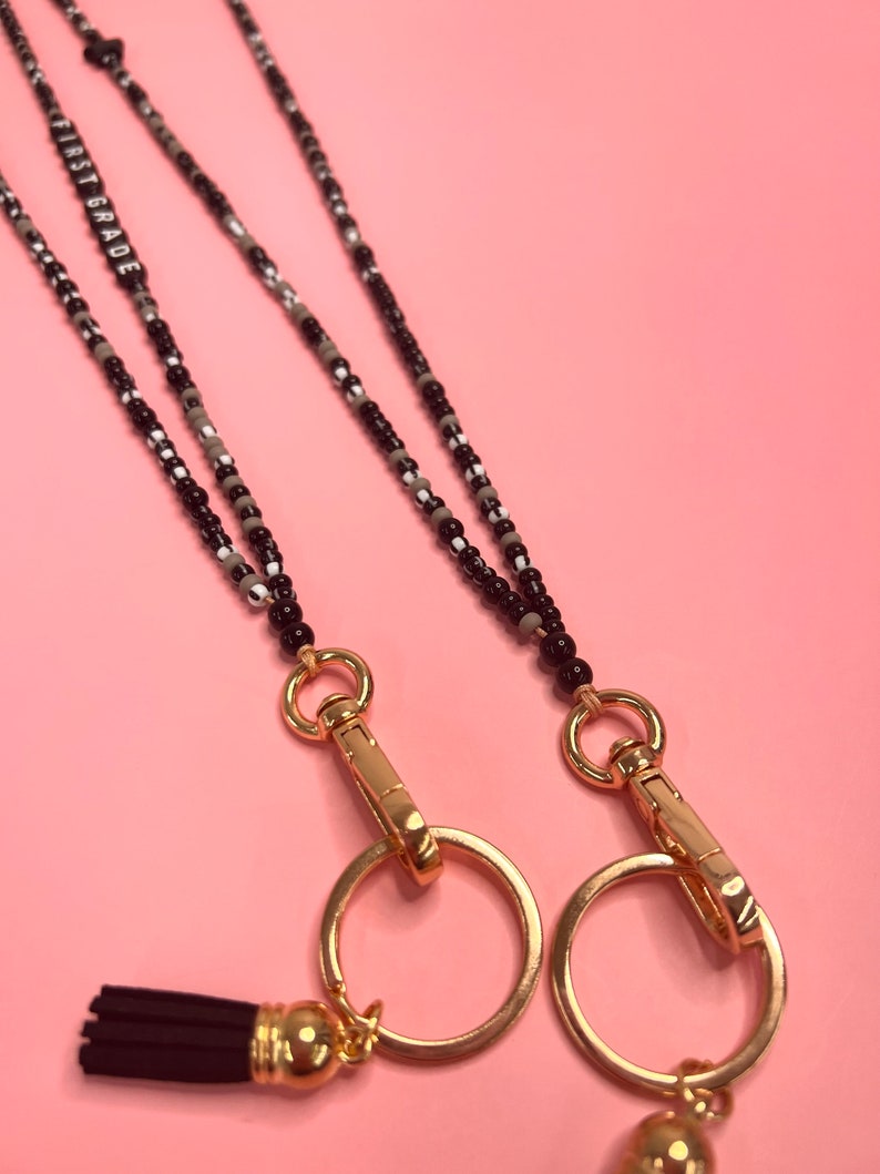 May include: Two black and white beaded lanyards with gold clasps and tassels. The lanyards have a gold charm with the text "FIRST GRADE" on it.
