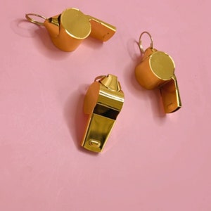 Peut inclure: Trois sifflets dorés sur un fond rose. Les sifflets ont un embout buccal rond et un petit trou sur le côté.