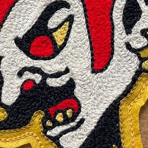 Puede incluir: Un parche bordado que muestra un rostro estilizado con piel blanca, detalles en negro y rojo. El dise&ntilde;o incluye formas amarillas similares a cuernos y un borde amarillo. El bordado utiliza hilos densos para crear una textura definida.