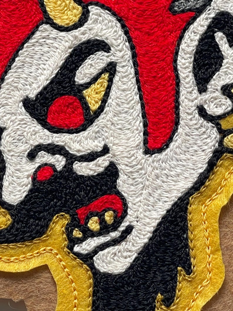 Puede incluir: Un parche bordado que muestra un rostro estilizado con piel blanca, detalles en negro y rojo. El dise&ntilde;o incluye formas amarillas similares a cuernos y un borde amarillo. El bordado utiliza hilos densos para crear una textura definida.