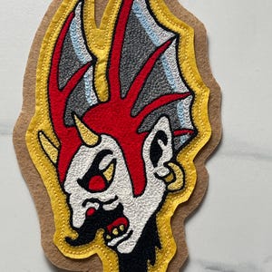 Puede incluir: Un parche bordado sobre una base de fieltro color canela que muestra una criatura estilizada de piel blanca, con una cresta roja brillante y cuernos afilados. El dise&ntilde;o incluye detalles en negro, gris y amarillo con un borde amarillo cosido.