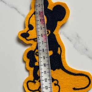 May include: A yellow felt patch featuring a black embroidered silhouette of a cartoon mouse. A white measuring tape is placed vertically over the patch, indicating a total height of approximately 15 centimeters or 6 inches from top to bottom.