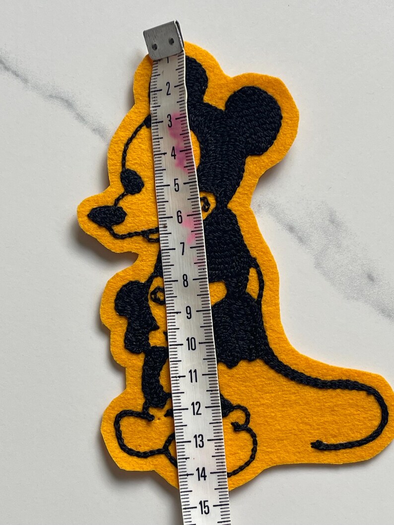 May include: A yellow felt patch featuring a black embroidered silhouette of a cartoon mouse. A white measuring tape is placed vertically over the patch, indicating a total height of approximately 15 centimeters or 6 inches from top to bottom.